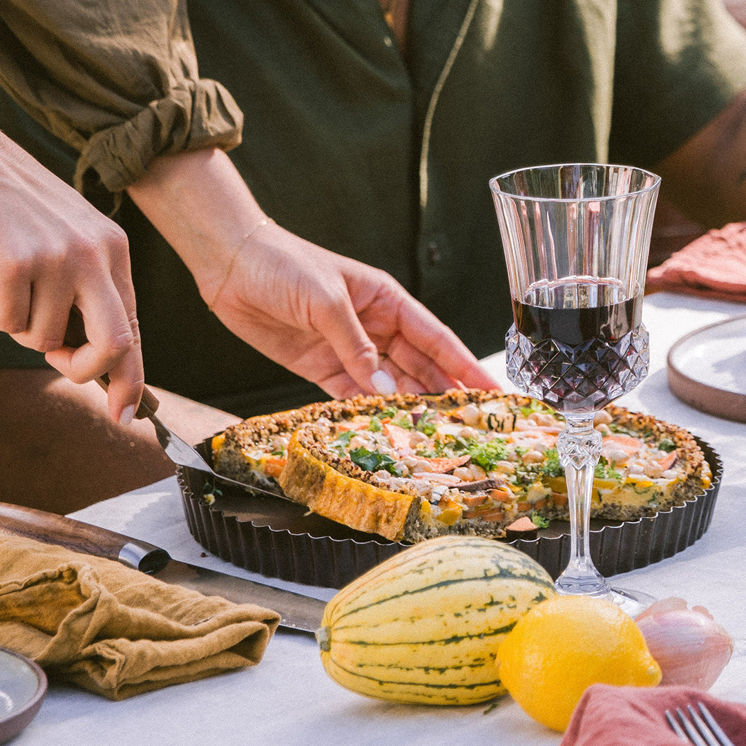Person serving a slice of homemade savory tart alongside a glass of red wine and fresh produce on a table.