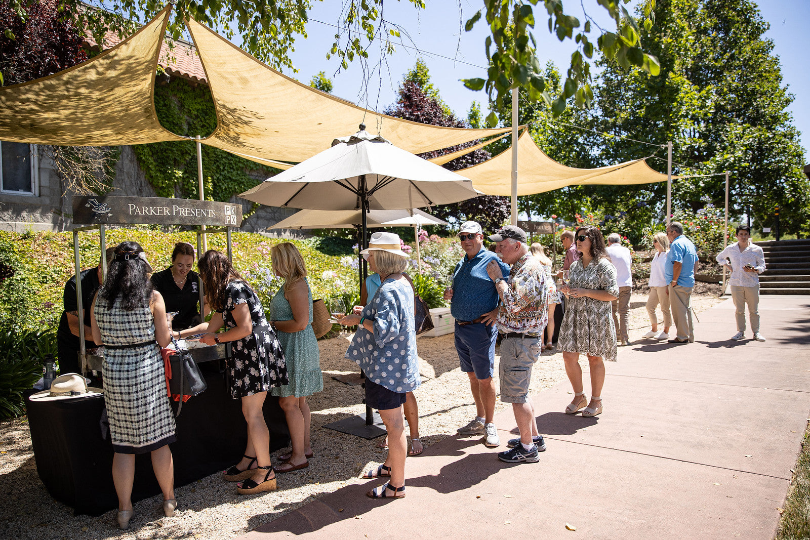 People line up for a glass of wine at an outdoor event with sun shades and umbrellas.