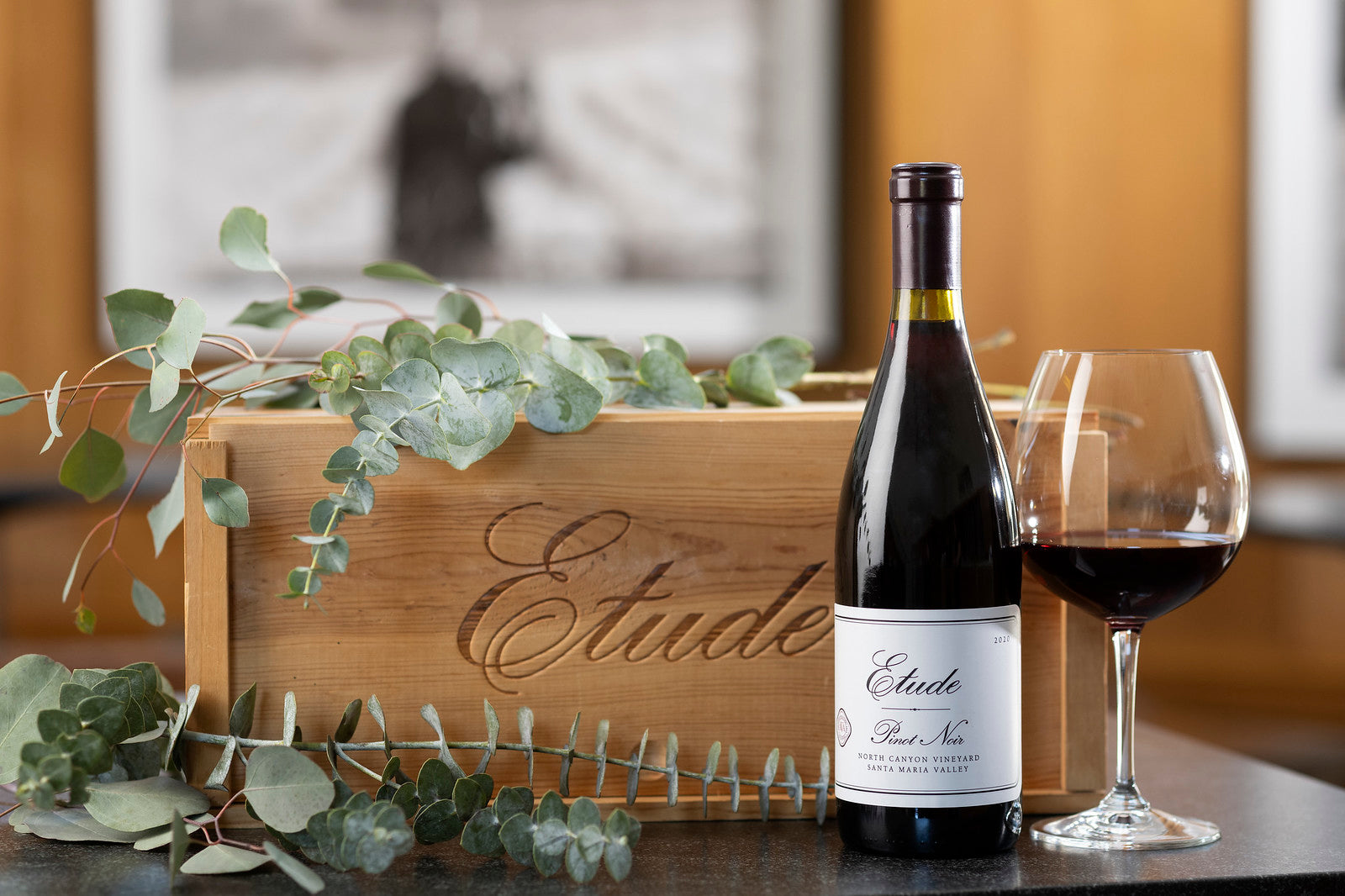 Etude Pinot Noir wine bottle and glass next to a wooden Etude box, eucalyptus leaves
