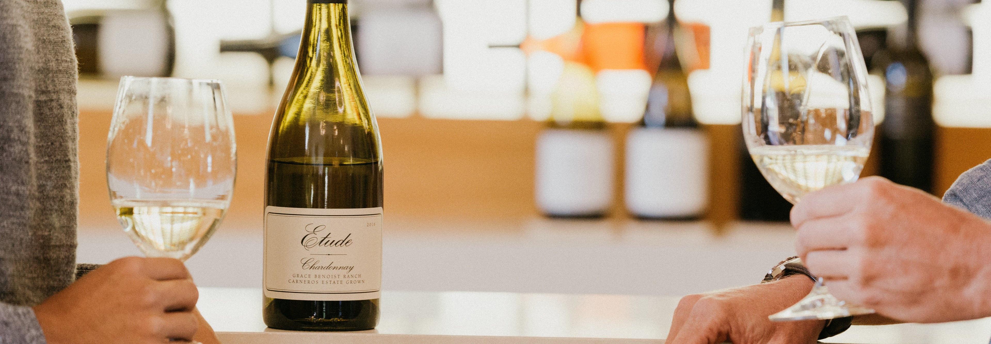 Two hands holding glasses of white wine, with a bottle of Etude Chardonnay in the background.